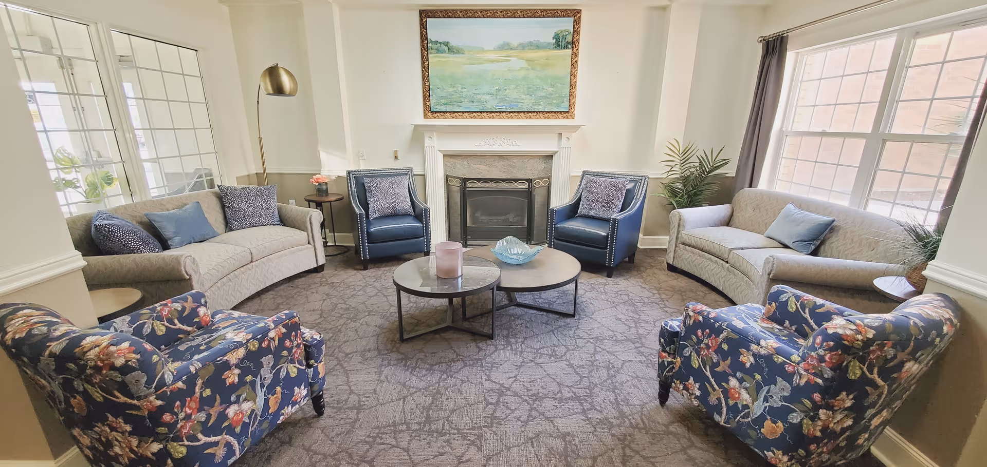 A cozy living room with two beige sofas, two blue armchairs, and two floral-patterned armchairs arranged around two round coffee tables. There is a fireplace with a landscape painting above it in the center of the room. Large windows on the right side let in natural light, and a tall floor lamp stands near one sofa.