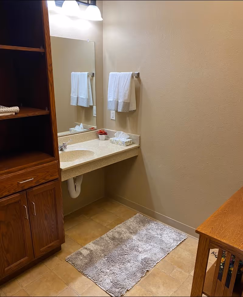 Bathroom vanity area with a sink, mirror, towel, tissue box, wooden cabinet and a gray bath mat on tiled floor.