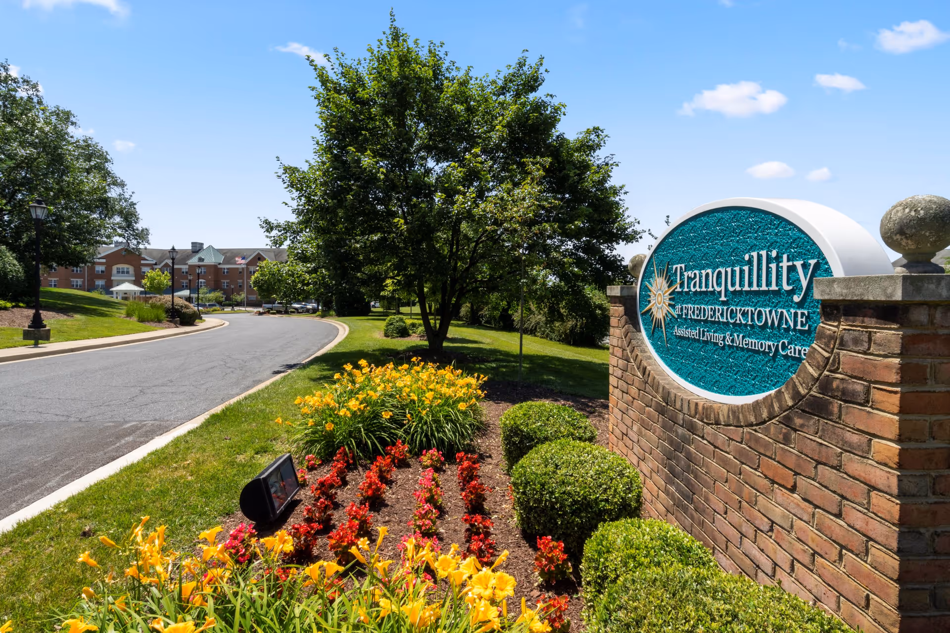 A landscaped entrance to Tranquillity at Fredericktowne assisted living and memory care facility, featuring a brick sign with the facility's name, colorful flower beds, trimmed bushes, a tree, and a paved road leading to the building under a clear blue sky.