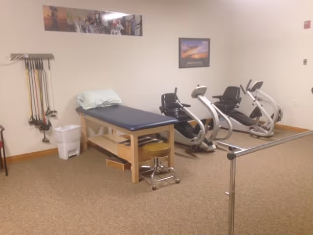 Therapy/fitness room with an examination table, two recumbent exercise bikes, resistance bands on the wall, and parallel bars.