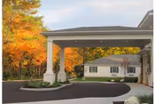 Entrance driveway of a senior living facility with a covered drop-off area, surrounded by autumn-colored trees and a white building in the background.