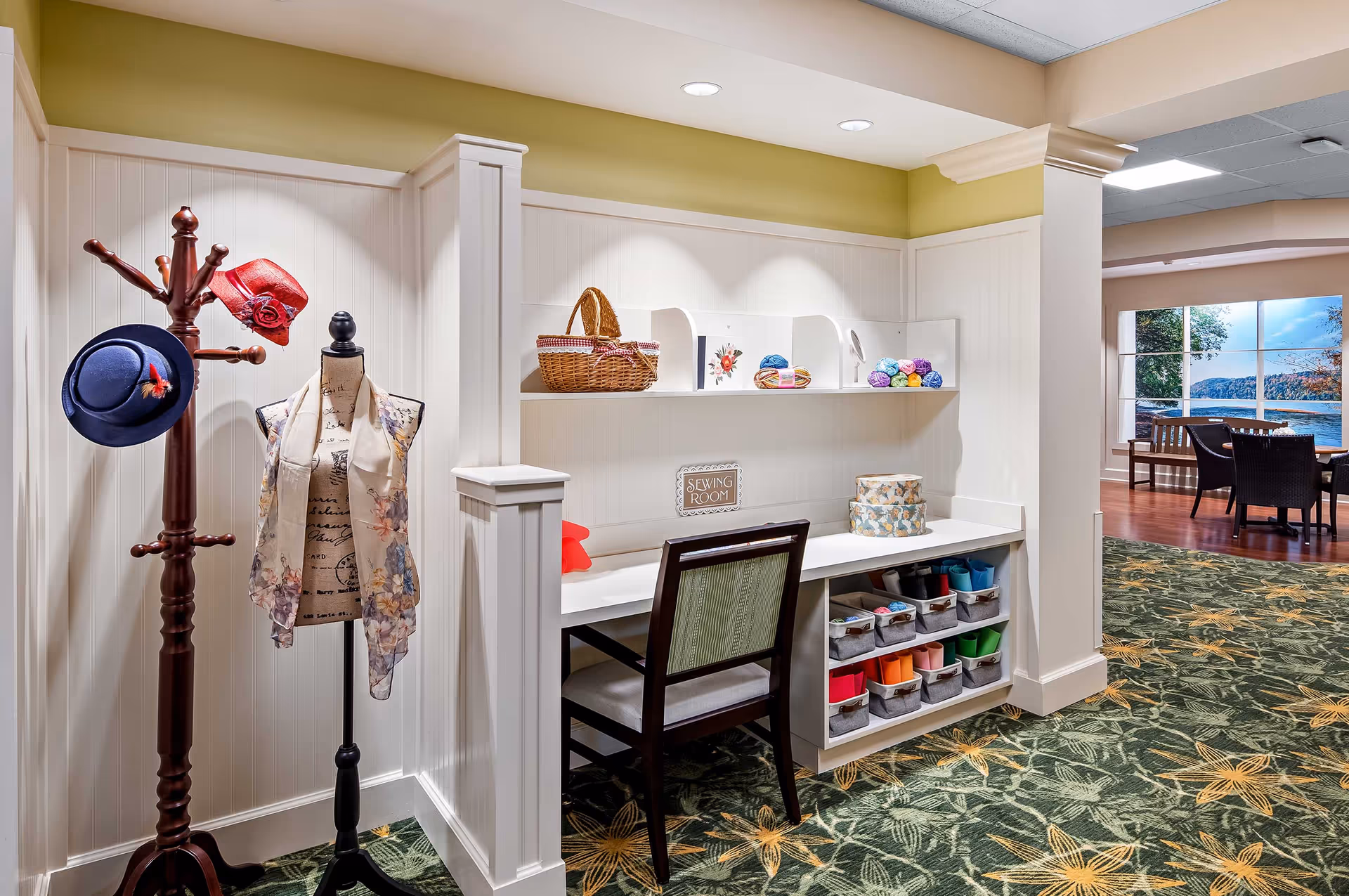 A cozy sewing room corner in a senior living facility with a wooden coat rack holding two hats, a dress form draped with a floral scarf, a desk with a chair, shelves holding yarn and baskets, and a sign that reads 'Sewing Room'. The area has white paneled walls, green and yellow floral carpet, and an adjacent seating area with a large window showing a scenic lake view.