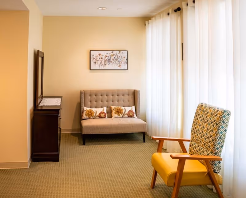 A small sitting area with a beige tufted loveseat adorned with three floral cushions, a patterned armchair with wooden armrests, a dark wooden dresser with a mirror, and sheer white curtains covering large windows, all set against light yellow walls and a green carpeted floor.