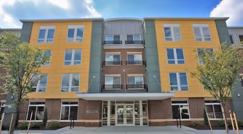 Front exterior view of Victory Square Apartments, a modern four-story building with a combination of yellow, green, and brick facade, large windows, and a central entrance with glass doors. There are small trees and a sidewalk in front of the building under a partly cloudy sky.