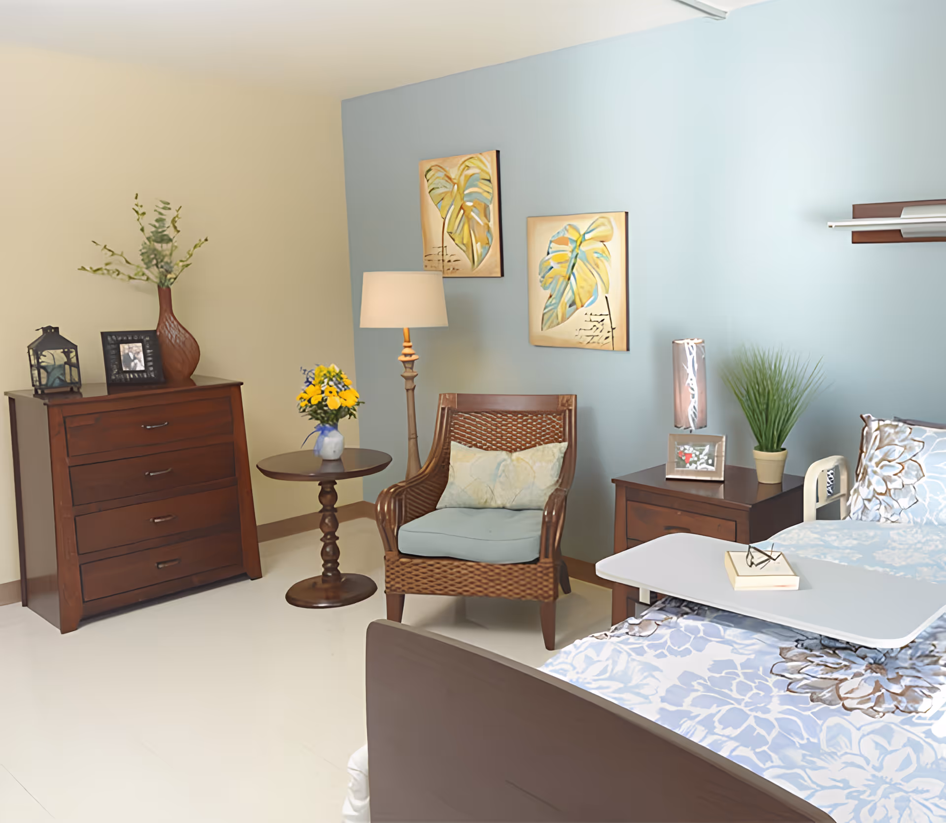 A cozy bedroom in a senior living facility featuring a bed with floral bedding, a wooden nightstand with a lamp and a potted plant, a wooden chair with a cushion, a small round table with a vase of flowers, a wooden dresser with decorative items, and two framed leaf paintings on a blue accent wall.