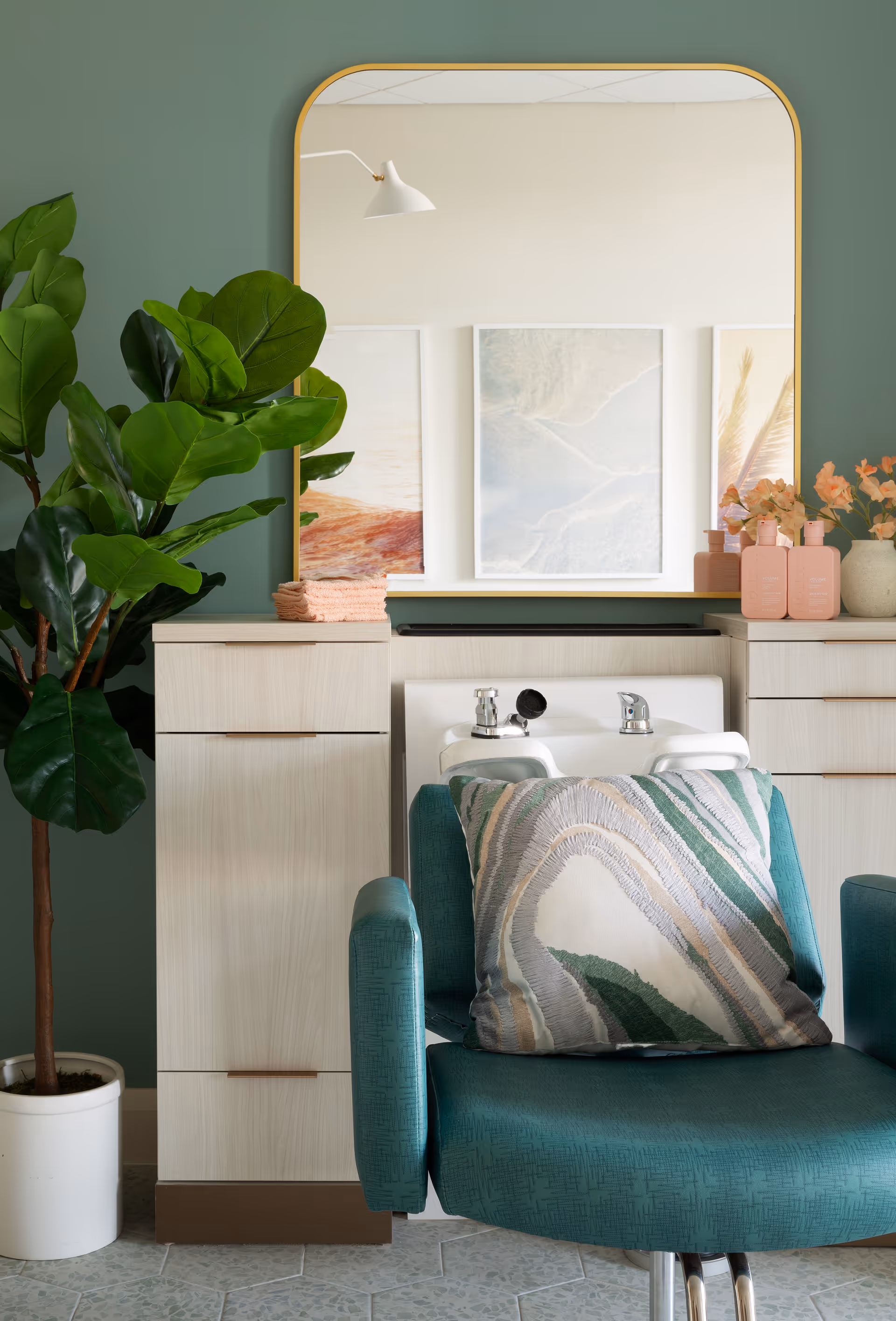 A teal salon chair with a decorative pillow in front of a white sink with two faucets. Behind the sink is a large mirror with a gold frame reflecting wall art and a white wall lamp. To the left of the chair is a tall green potted plant, and to the right are light-colored cabinets with pink bottles and flowers on top.