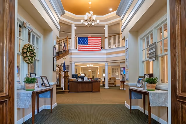 Interior view of a senior living facility lobby with a high ceiling, chandelier, American flag hanging on the upper balcony, wooden reception desk, and decorative plants on tables along the hallway.