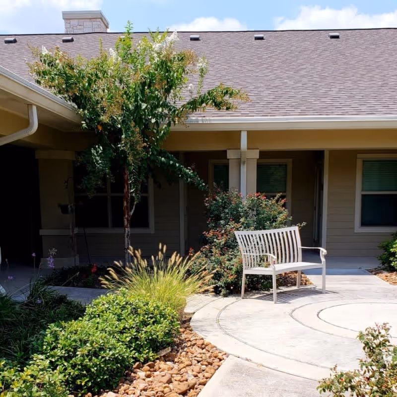 Outdoor courtyard area at Orchard Park at Southfork Assisted & Senior Living featuring a white metal bench on a circular concrete pathway surrounded by green shrubs, ornamental grasses, a small tree, and flowering plants under a clear blue sky.