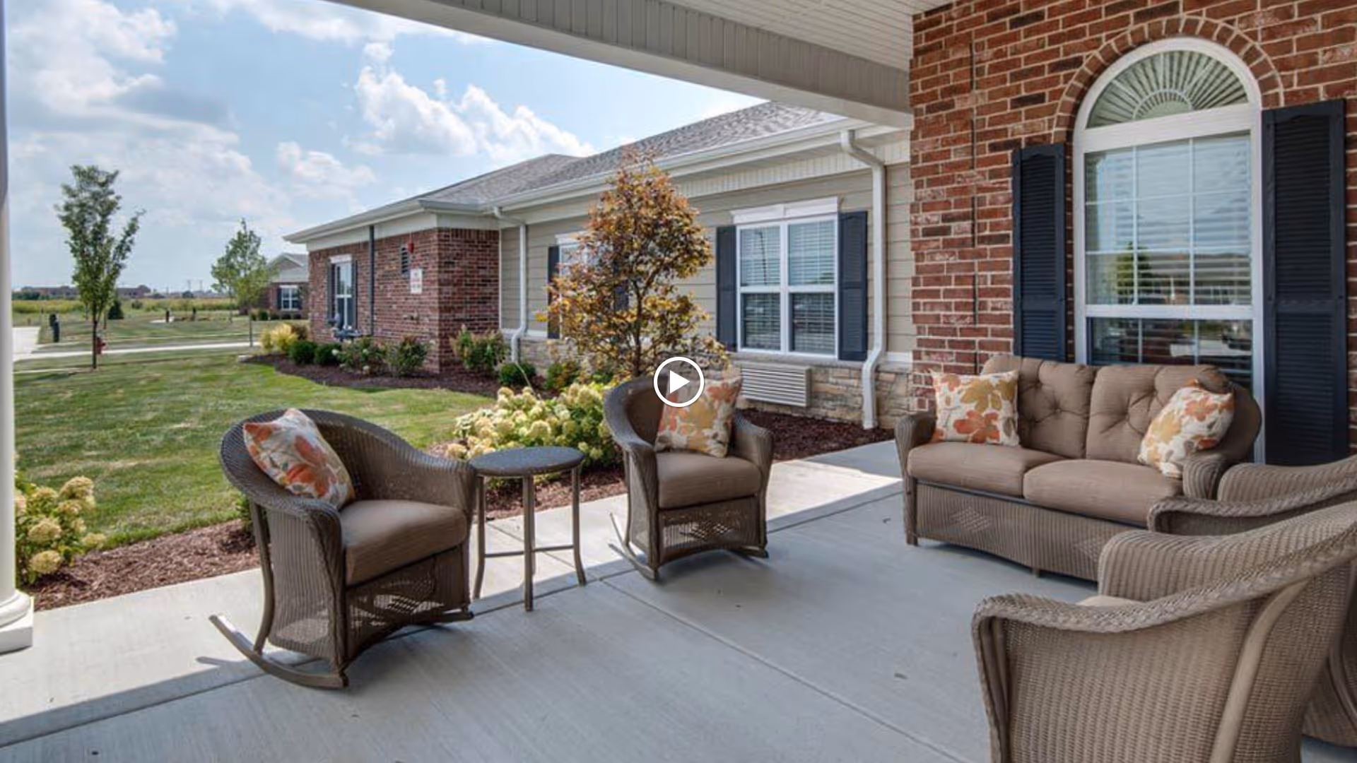 Outdoor patio area at Mattis Pointe Senior Living with wicker furniture including a sofa, two armchairs, and a rocking chair, all with floral cushions. The patio is adjacent to a brick building with windows and black shutters, surrounded by landscaped greenery and a clear sky.
