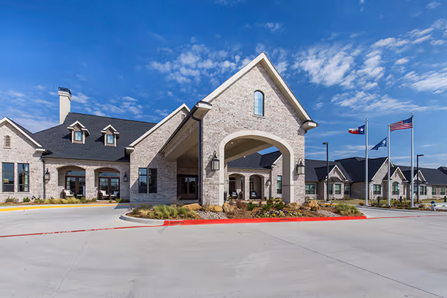 Exterior view of a senior living facility building with a covered entrance, brick facade, multiple windows, and three flagpoles displaying the Texas state flag, the American flag, and another flag. The sky is clear with some clouds.