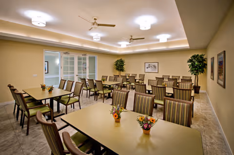 A well-lit dining room with multiple rectangular tables and striped cushioned chairs arranged neatly. Small flower arrangements are placed on the tables. The room has beige walls, tiled floors, ceiling fans, and recessed lighting. There are framed pictures on the walls and potted plants in the corners.