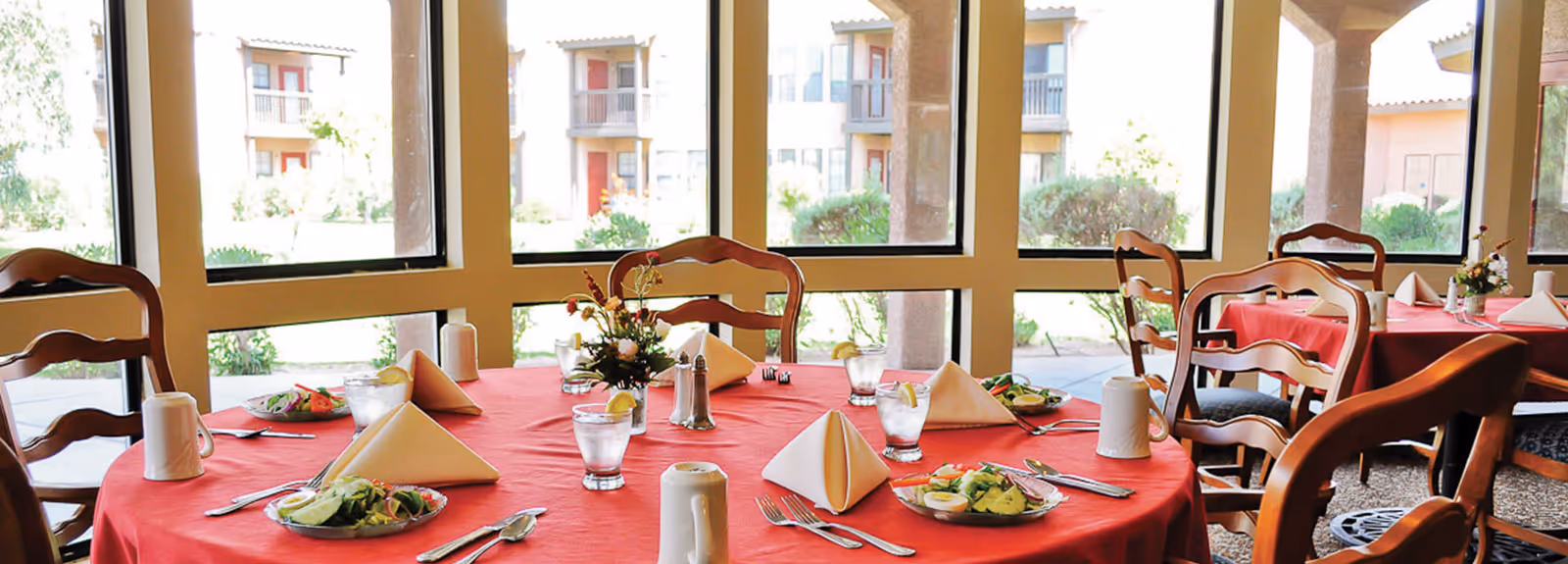 Dining area with round tables covered in red tablecloths, set with plates of salad, glasses of water with lemon, folded napkins, and silverware. Wooden chairs surround the tables. Large windows in the background provide a view of the outdoor garden and building exterior.