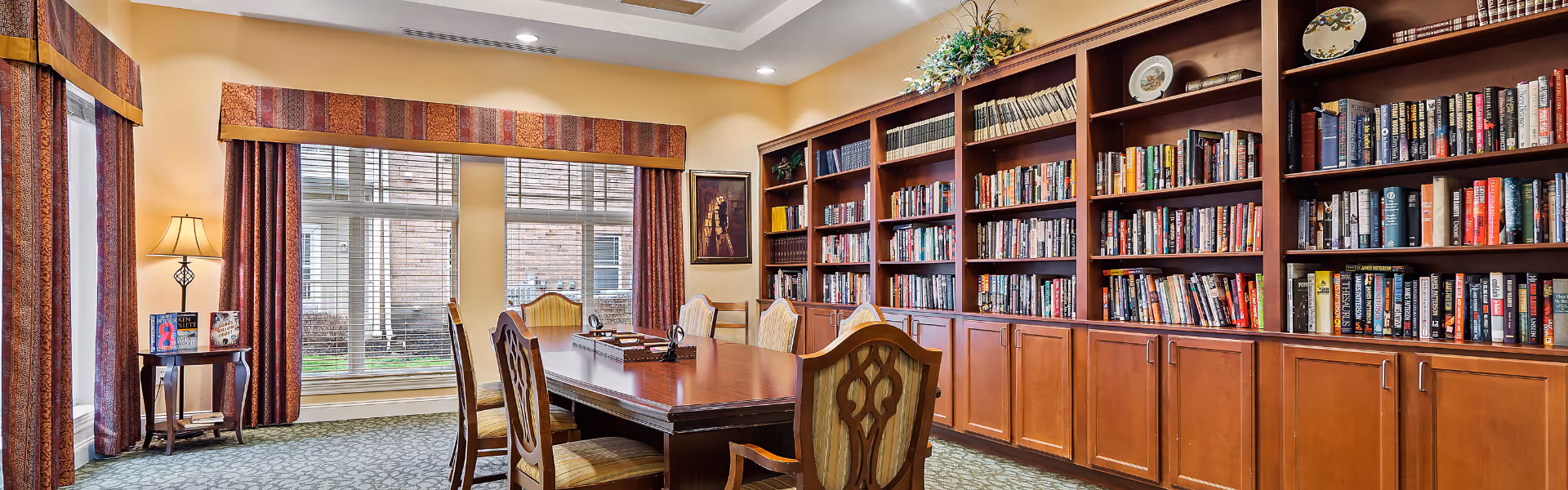 A cozy library or reading room with a large wooden table surrounded by six upholstered chairs. The room features built-in wooden bookshelves filled with books and decorative plates. Large windows with blinds and patterned valances let in natural light, and a small side table with a lamp and books is positioned near the window.