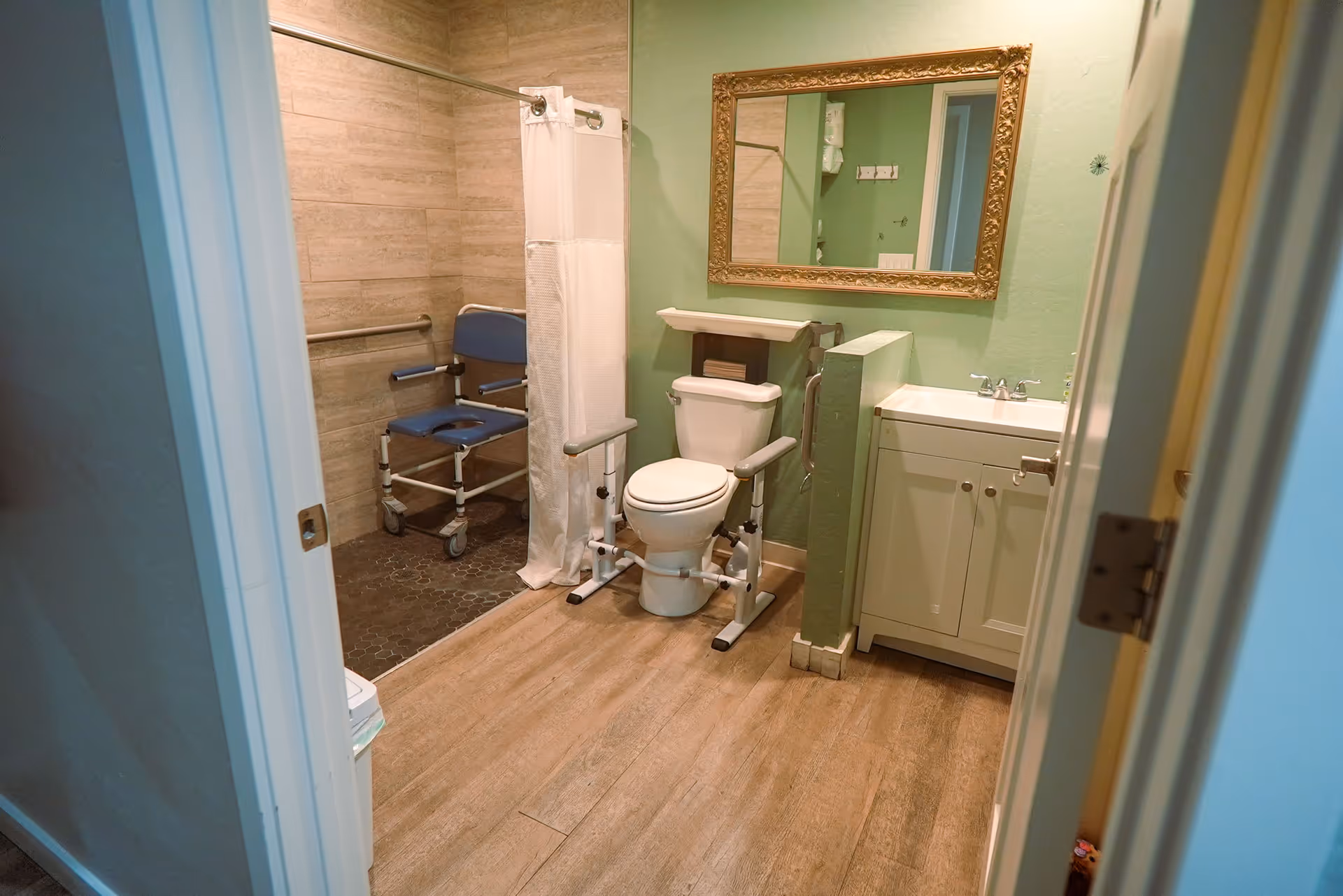 A bathroom with a green wall, a toilet equipped with safety rails, a white vanity with a sink, a large ornate mirror above the sink, and a shower area with a white curtain and a blue shower chair with wheels.