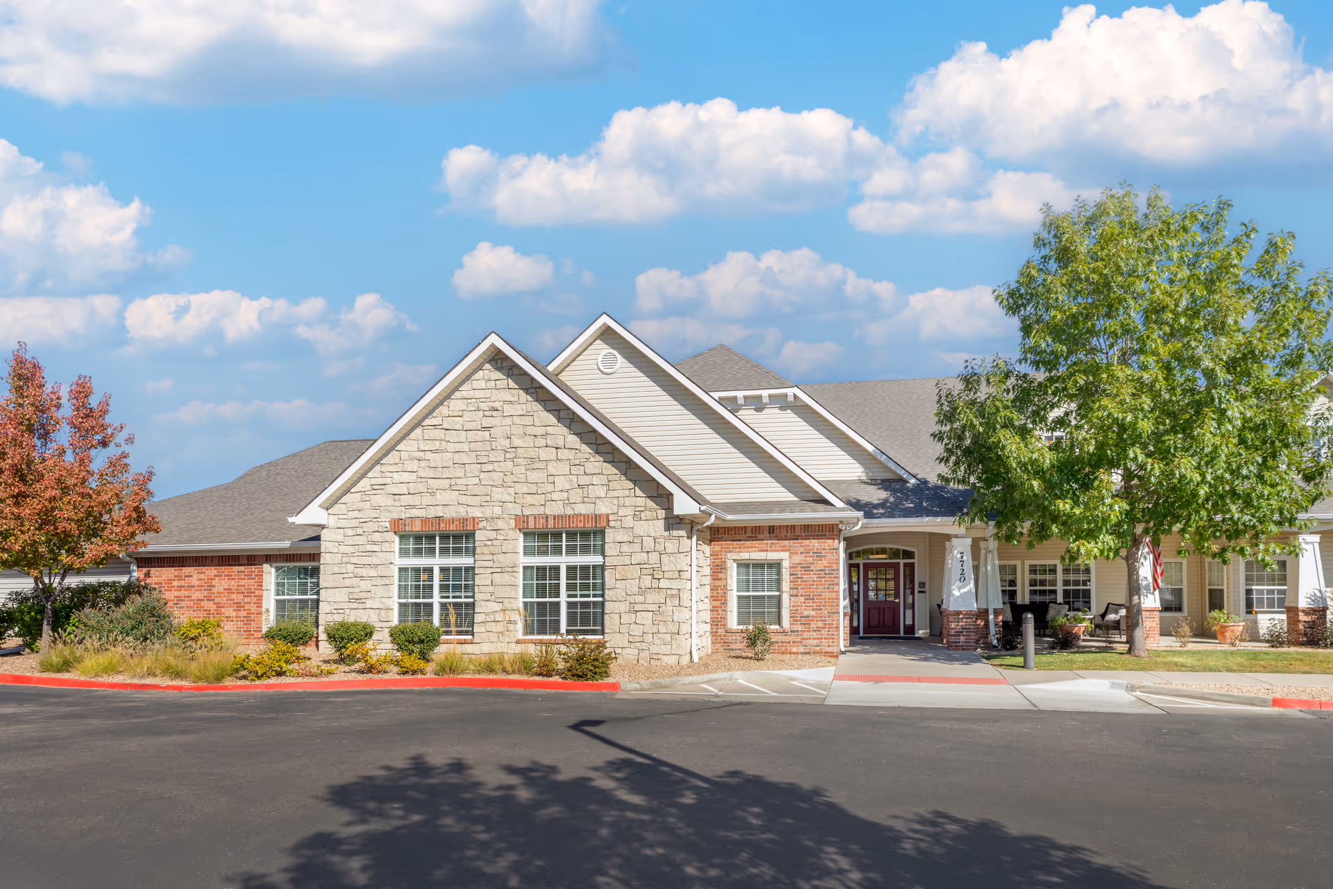 Exterior view of a single-story senior living facility building with a combination of stone and brick facade, multiple windows, a covered entrance with a maroon door, and surrounding landscaping including trees and shrubs under a partly cloudy blue sky.