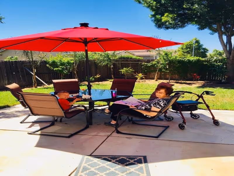 An elderly woman and a young child sitting outside on patio chairs around a glass table under a large red umbrella. The elderly woman is reading a newspaper and a red walker is nearby. The background shows a fenced garden with green grass, plants, and trees under a clear blue sky.