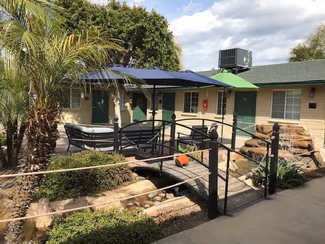 Outdoor courtyard area at Arizona Desert Falls featuring a small wooden bridge over a narrow water feature with rocks, surrounded by greenery including palm trees and bushes. There are patio tables with blue and green umbrellas, and a beige building with green doors and windows in the background under a partly cloudy sky.