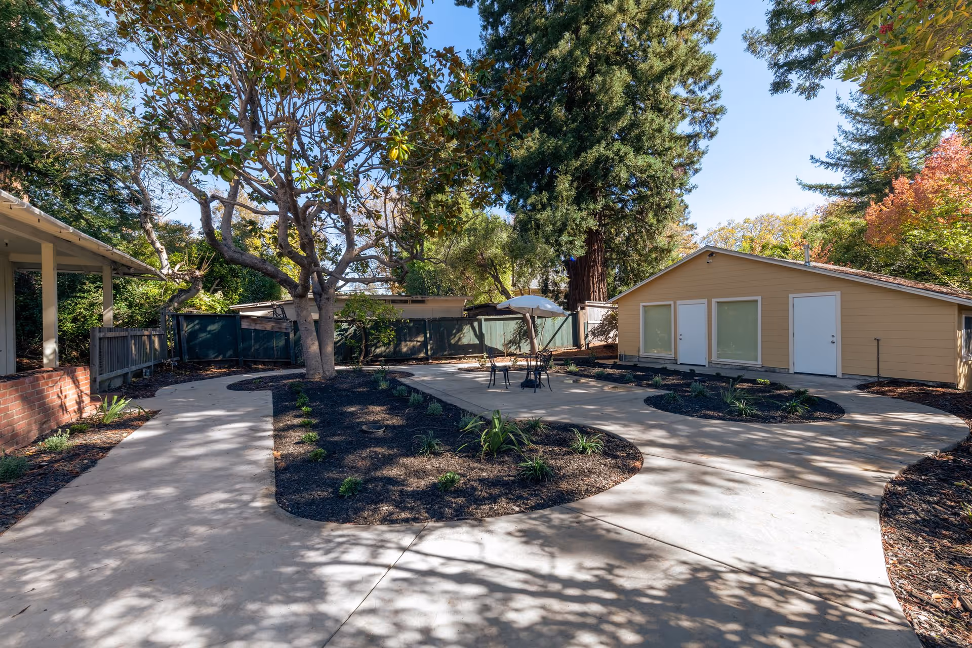 Outdoor area at Marin Terrace Assisted Living featuring a paved walkway curving around landscaped garden beds with small plants and a large tree in the center. There is a small patio with a table, chairs, and an umbrella. A beige building with three doors and windows is visible in the background, surrounded by tall trees and greenery under a clear blue sky.