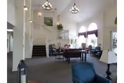 Spacious and well-lit living room area in an assisted living facility featuring multiple seating arrangements with chairs and tables, large windows with arched tops allowing natural light, a staircase leading to an upper floor, and decorative light fixtures hanging from the ceiling.