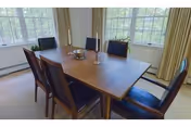 A dining room with a rectangular wooden table surrounded by six dark wooden chairs with black cushions. The room has large windows with beige curtains, allowing natural light to fill the space. On the table, there are two candlesticks and a small decorative bowl.