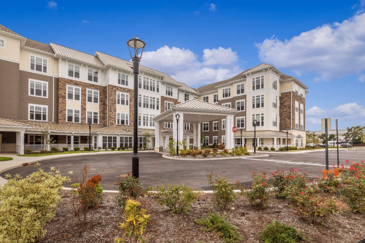 Front exterior of a multi-story senior living facility with a covered entrance, circular driveway, landscaping, and lampposts under a blue sky.