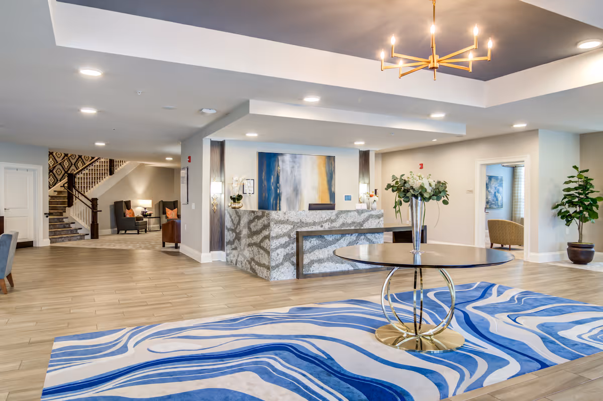 Bright and spacious senior living facility lobby with a modern reception desk made of marble, a round table with a vase of flowers on a blue and white patterned rug, comfortable seating areas, potted plants, and a staircase leading to the upper floor.