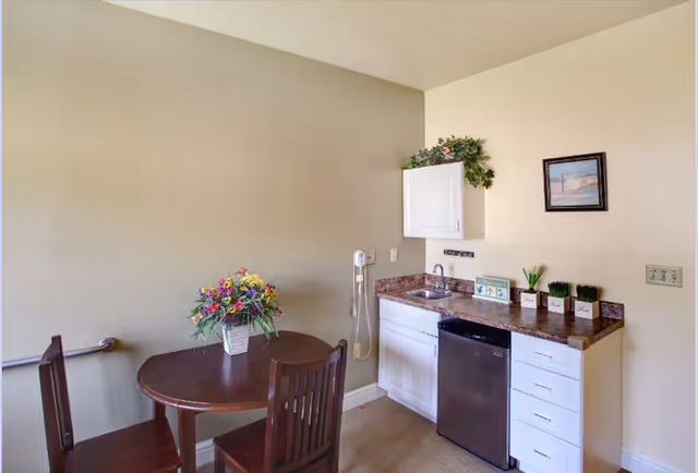 Small kitchenette area with a round wooden table and two chairs. A vase with colorful flowers is on the table. The kitchenette has a countertop with a sink, a small black refrigerator, white cabinets, and some decorative plants on the counter. A wall-mounted phone and a framed picture are also visible on the beige walls.