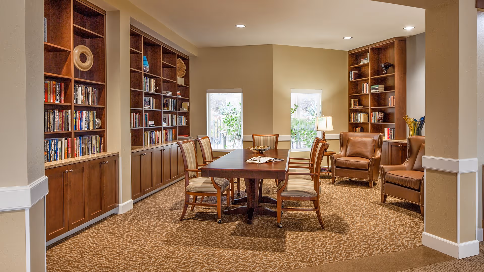 A cozy library or reading room with built-in wooden bookshelves filled with books, a rectangular wooden table surrounded by six cushioned chairs, and three brown leather armchairs near two small windows with natural light. The room has beige walls and patterned carpet flooring.