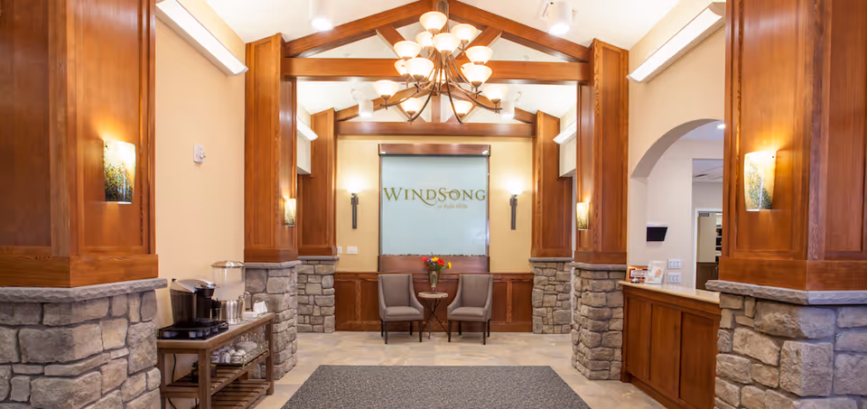 Interior view of a senior living facility lobby with stone and wood pillars, a chandelier hanging from a wooden beam ceiling, two chairs and a small table with flowers in front of a frosted glass panel displaying the text 'Windsong at Eola Hills'.