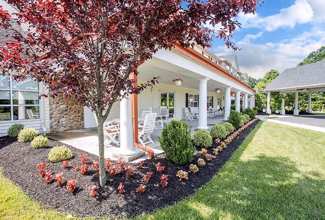 A bright outdoor view of a senior living facility porch with white columns, rocking chairs, hanging flower pots, and a landscaped garden bed with small bushes and red-leafed tree under a partly cloudy sky.