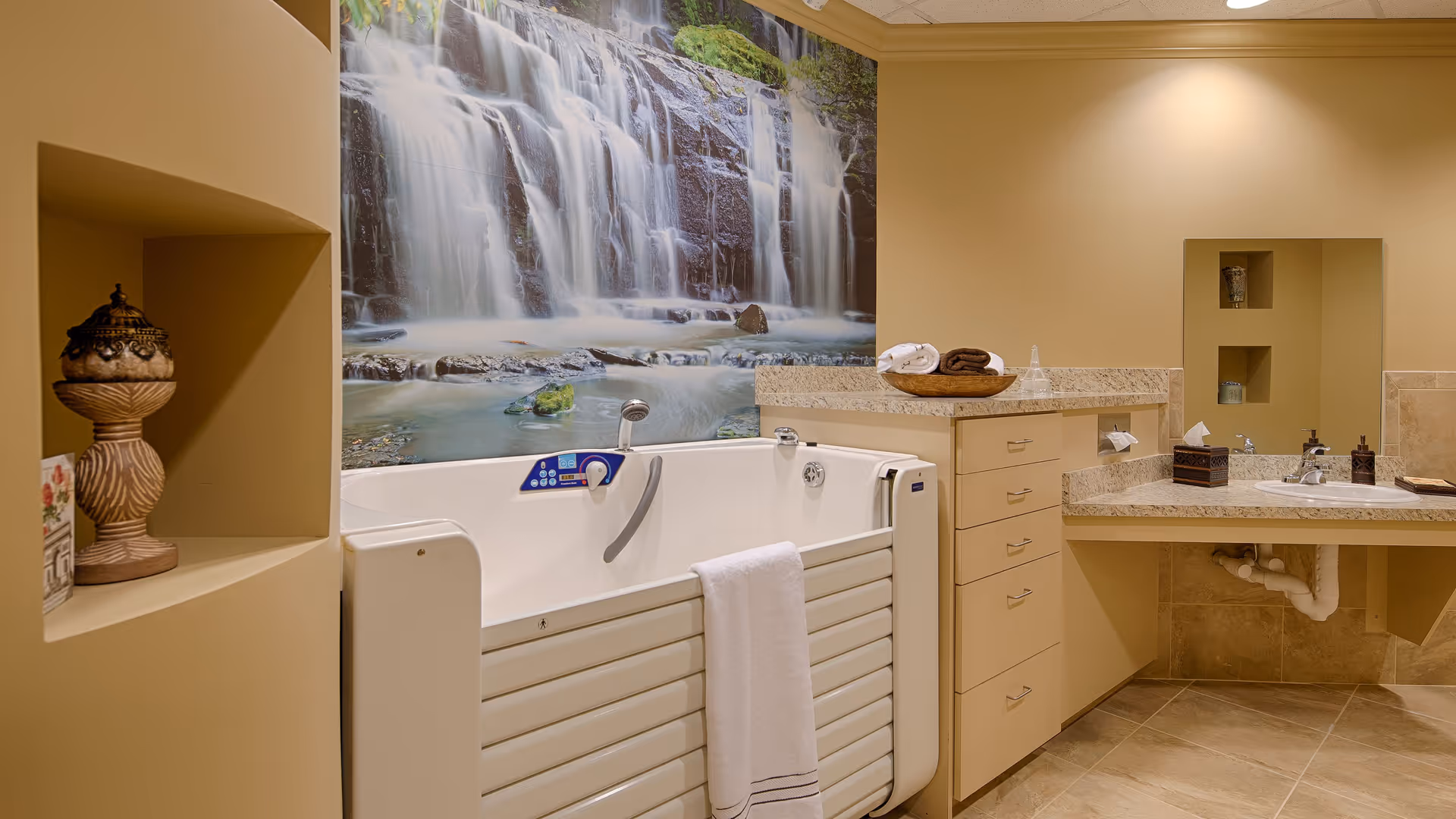 A bathroom with a walk-in bathtub featuring a control panel and a grab bar. A white towel hangs on the tub. The walls are beige, and there is a large mural of a waterfall above the bathtub. The bathroom also has a countertop with drawers, a sink, a mirror, and decorative items including a basket with towels and a small vase.