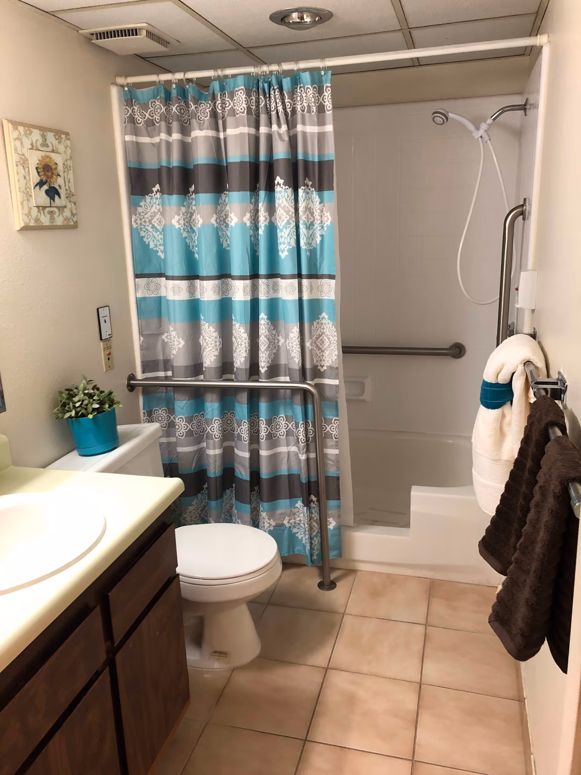 Accessible bathroom with a toilet, vanity sink, tiled floor, and a bathtub-shower with a blue-and-gray patterned curtain and grab bars.