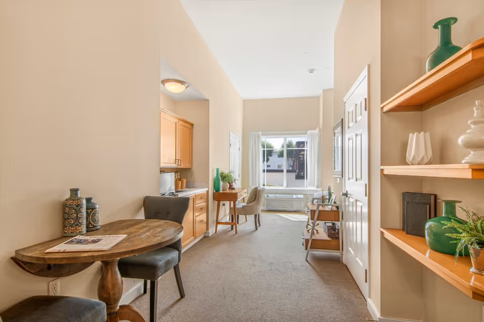A bright, narrow living space with beige walls and carpeted floor. On the left, there is a small wooden table with two chairs and decorative vases. Further back, a kitchenette with wooden cabinets is visible. At the end of the room, a window with white curtains lets in natural light, illuminating a desk and chair. On the right side, wooden shelves hold decorative items and a door is visible.