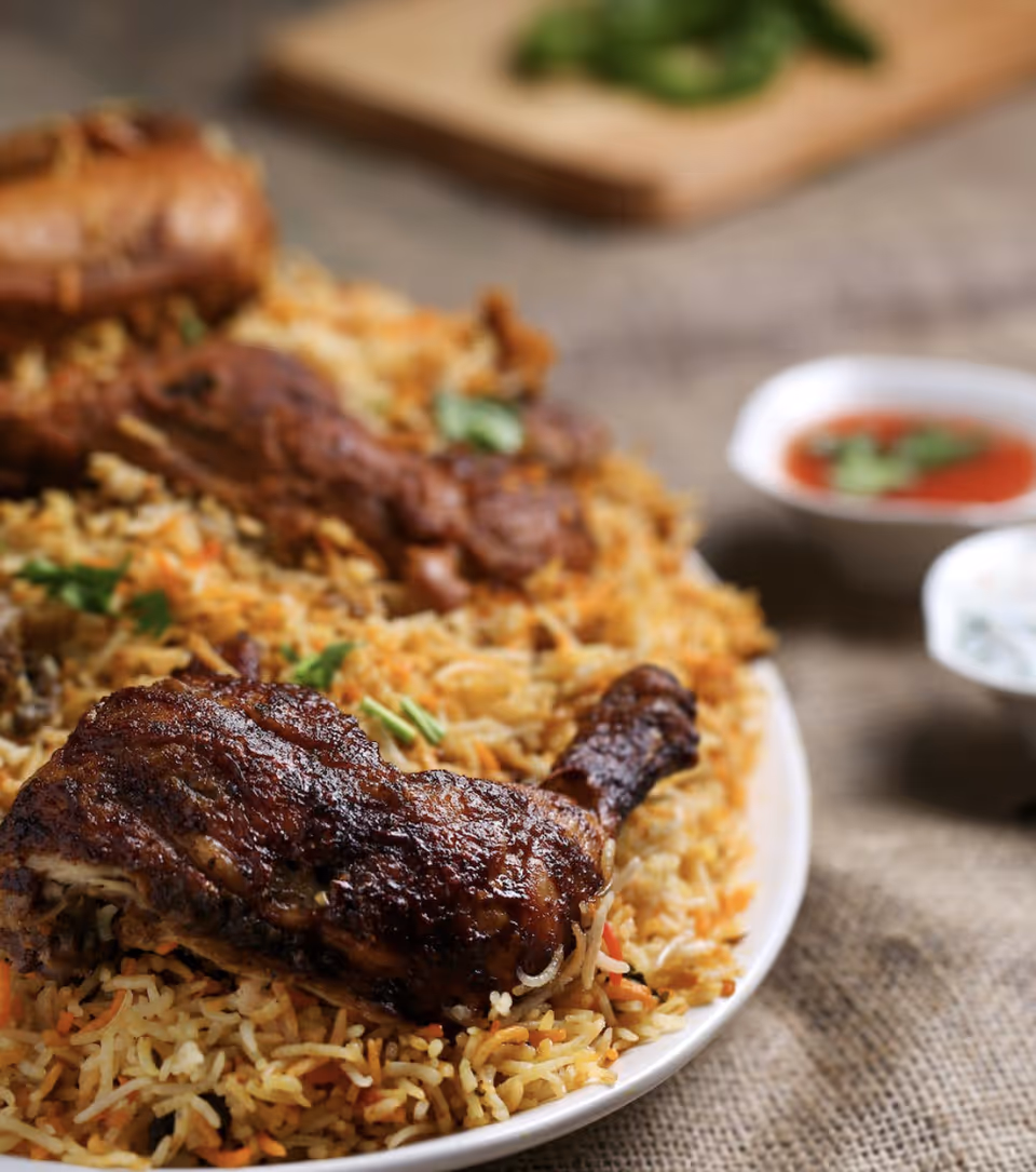 Close-up of a plate with seasoned grilled chicken drumsticks served on a bed of spiced rice, with small bowls of red sauce and white sauce in the background.