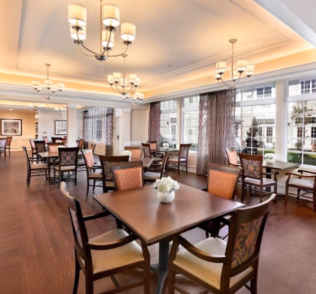 A spacious and well-lit dining room with multiple wooden tables and chairs arranged neatly. Each table has a small white floral centerpiece. Large windows with sheer curtains allow natural light to fill the room. Elegant chandeliers hang from the ceiling, enhancing the warm and inviting atmosphere.