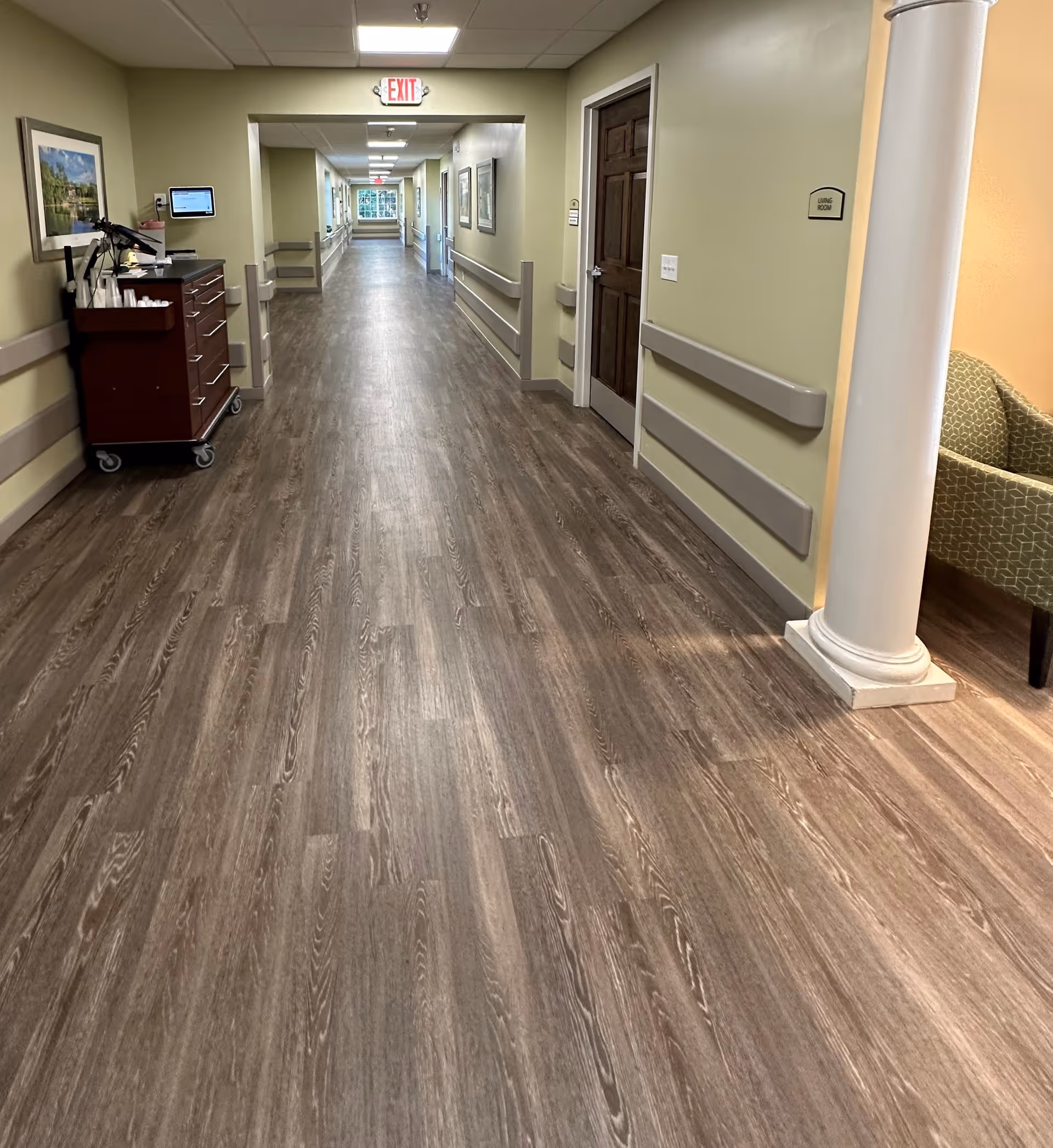 Long, well-lit interior hallway with wood-look flooring, handrails, an exit sign, and a service cart.