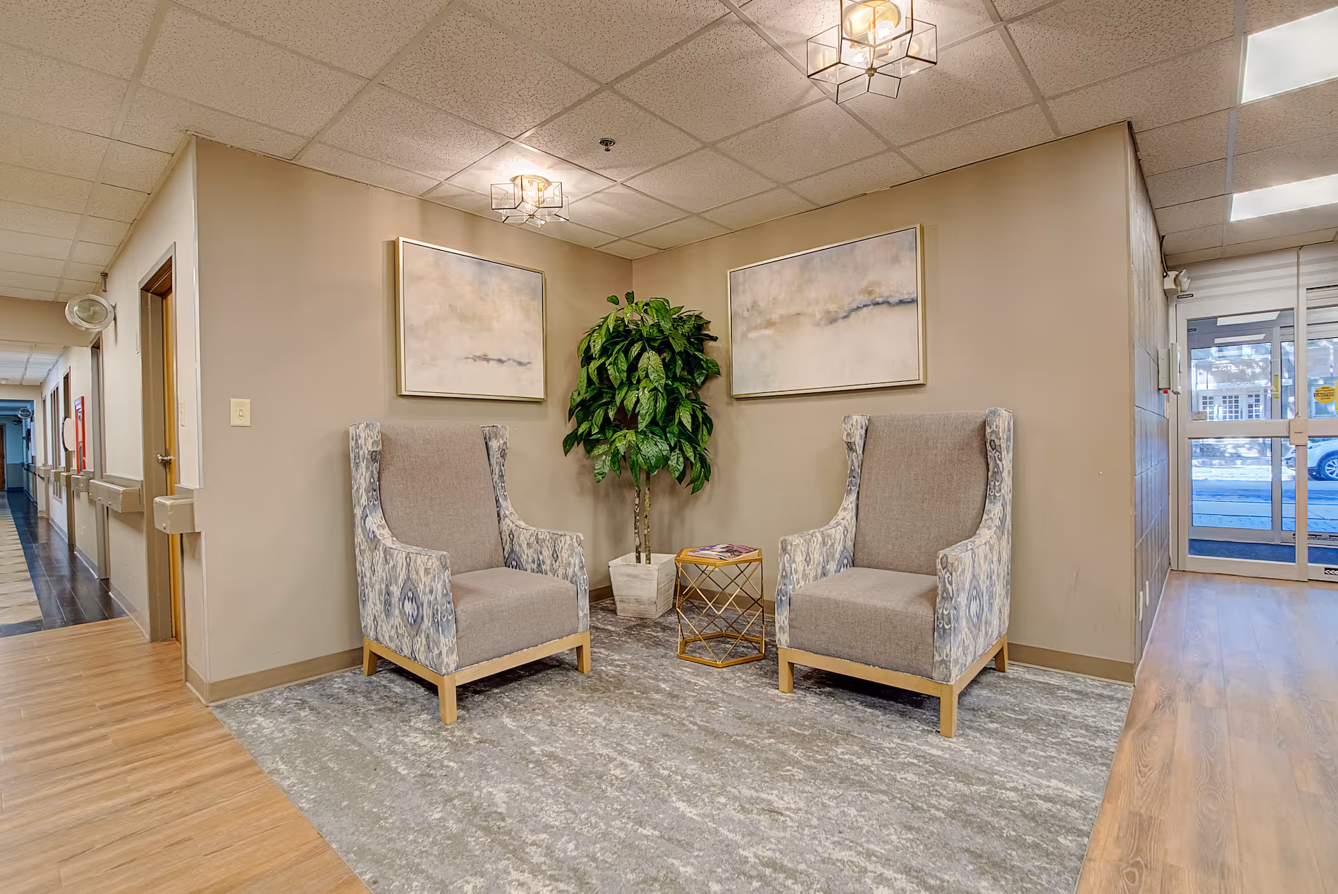 A small seating area in a hallway with two patterned armchairs facing each other, a small round gold side table between them, a potted green plant behind the table, and two abstract paintings on the beige walls. The floor is covered with a gray patterned rug, and the hallway has light wood flooring and a ceiling with recessed lighting. A glass door leading outside is visible in the background.