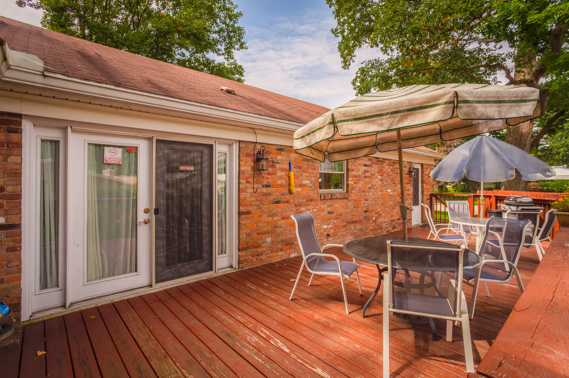 Outdoor wooden deck area with a round glass table and several chairs. Two large umbrellas provide shade. The deck is attached to a brick building with a sliding glass door and a screen door. Trees and a red wooden fence are visible in the background.