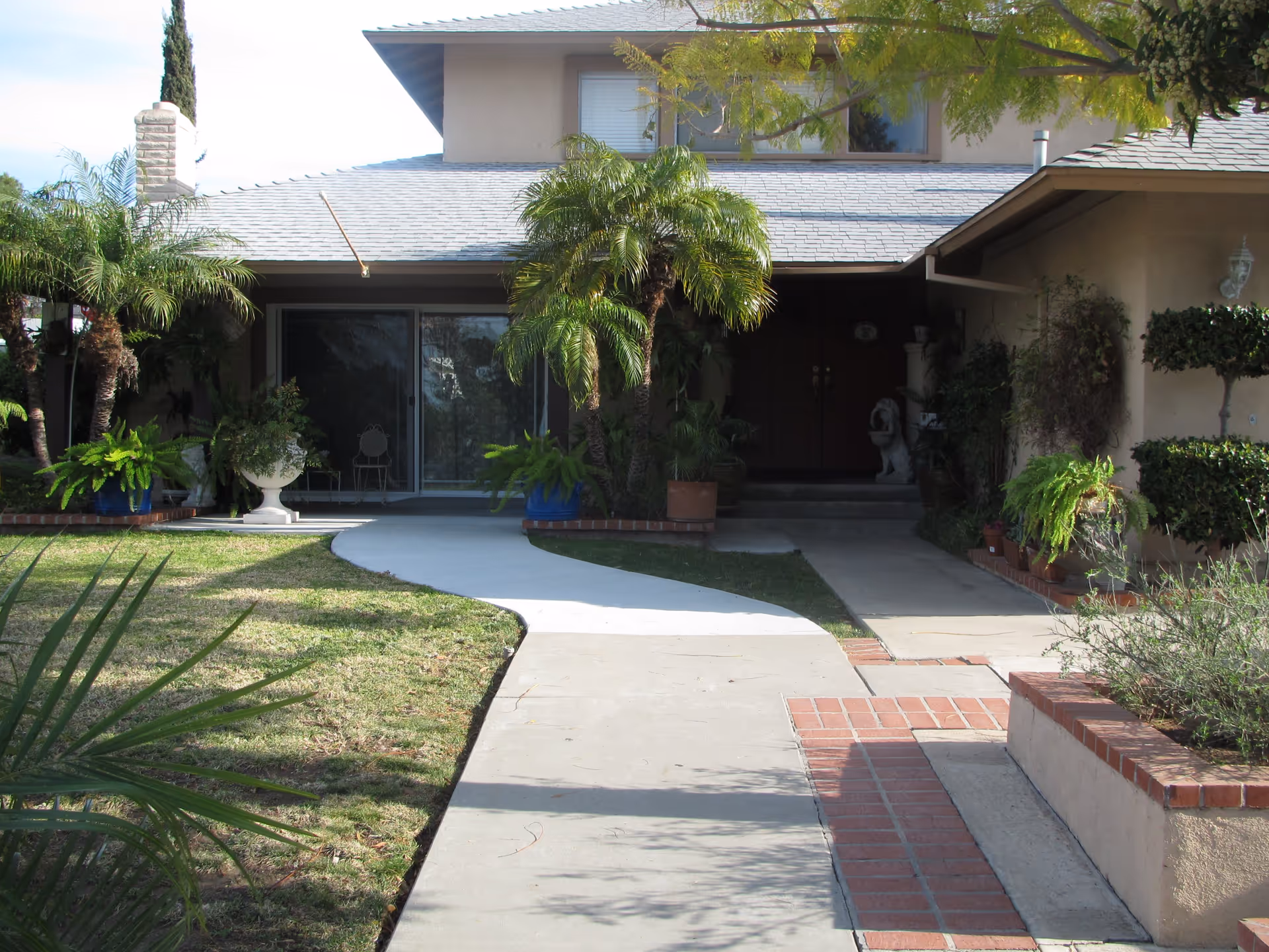 Front exterior view of a two-story house with a gray shingled roof, a curved concrete walkway leading to the entrance, surrounded by green grass, palm trees, and various potted plants.