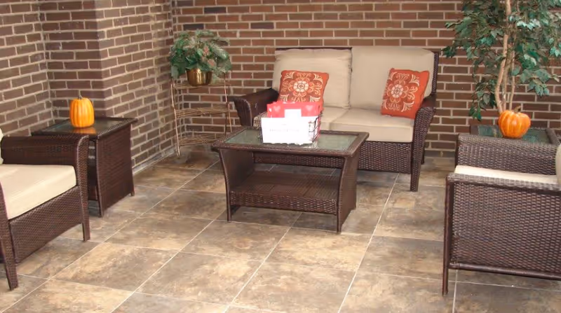 A cozy indoor seating area with wicker furniture including a loveseat and two chairs with beige cushions and orange floral pillows. There is a glass-top coffee table and two side tables, each decorated with a small pumpkin. The walls are made of brown brick and there are two potted plants in the corners.