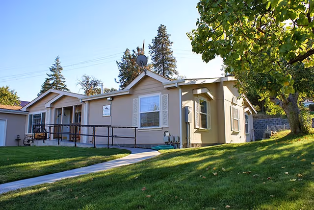 Single-story beige senior care building front with an accessibility ramp, green lawn, and trees under a clear sky.