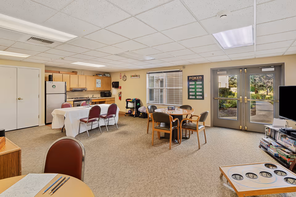 A spacious common area in a senior living facility featuring a kitchenette with wooden cabinets, a refrigerator, microwave, and stove. There are two tables with chairs, one covered with a white tablecloth and the other round with four chairs. The room has beige walls, a carpeted floor, and a ceiling with fluorescent lights. Double glass doors lead outside to a garden area. Various games and recreational items are visible, including a poker score chart on the wall and board games on a shelf near a television.