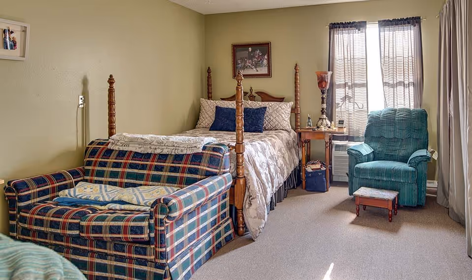 A cozy bedroom with a wooden four-poster bed adorned with beige and blue pillows and a patterned comforter. In front of the bed is a plaid loveseat with folded blankets on it. To the right, there is a green upholstered armchair with a small wooden footstool. A window with sheer curtains allows natural light into the room. A wooden side table next to the bed holds a tall decorative lamp and other small items. The walls are painted light green, and a framed picture hangs above the bed.