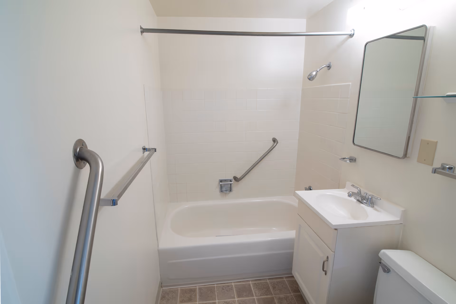 Small accessible bathroom with a bathtub/shower, grab bars, vanity sink, mirror, and toilet.