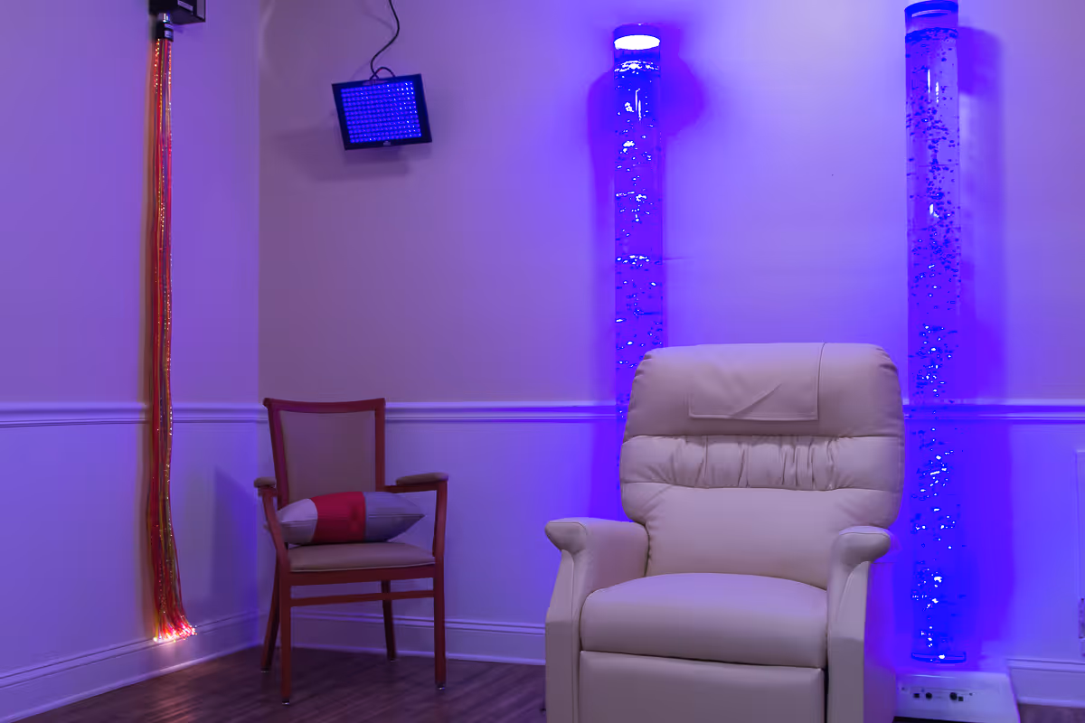 A softly lit room with a beige recliner chair and a wooden chair with a cushion. The room features two tall bubble tubes emitting blue light, a fiber optic light strand hanging on the wall, and a small square blue light fixture mounted on the wall. The walls are painted light purple with white trim, and the floor is wooden.