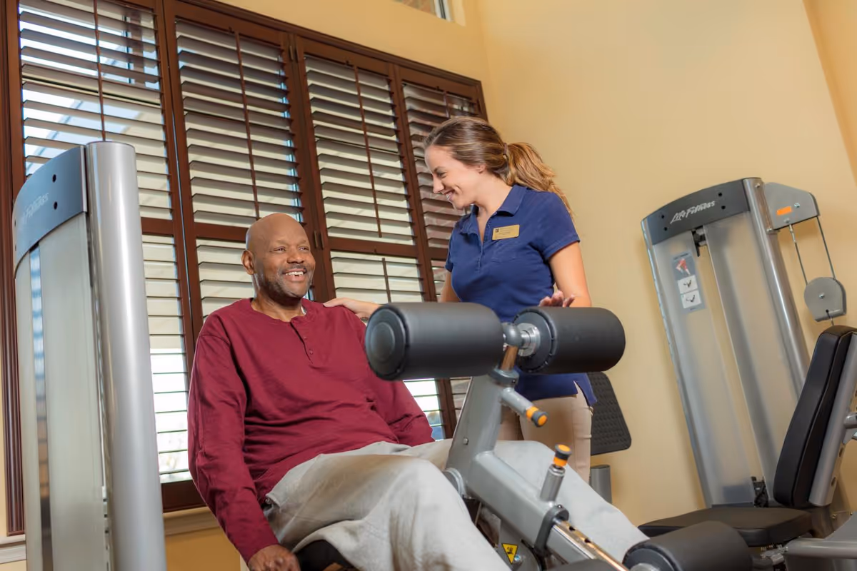 A man in a maroon long-sleeve shirt and gray pants is seated on a leg exercise machine in a fitness room, smiling and looking at a woman standing beside him. The woman, wearing a navy blue polo shirt and beige pants, is smiling and has her hand on the man's shoulder. The room has large windows with wooden blinds and additional exercise equipment.