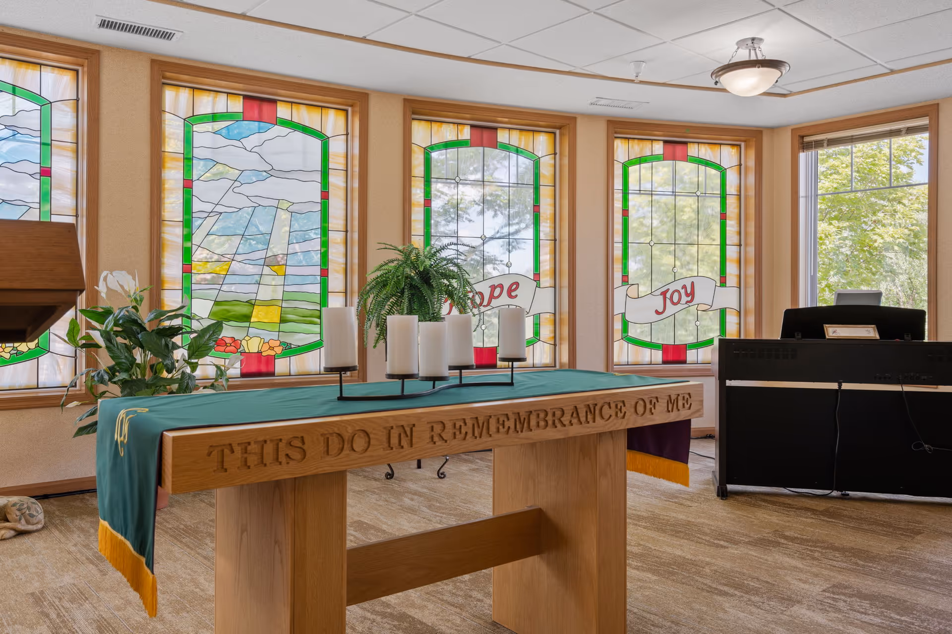 Sunlit chapel interior with a wooden communion table engraved "THIS DO IN REMEMBRANCE OF ME", candles, plants and stained-glass windows reading "Hope" and "Joy".