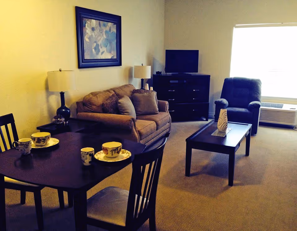 A cozy living room in an assisted living facility featuring a brown sofa with cushions, a dark wooden coffee table with a decorative vase, a dark armchair near a large window with blinds, a dark wooden dining table set with cups and saucers, two table lamps, a framed floral painting on the wall, and a dark wooden dresser with a flat-screen TV on top.
