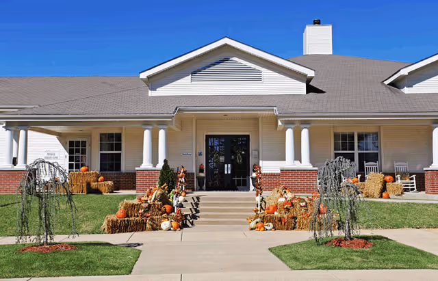 Front exterior view of a single-story building with white siding and a gray roof, decorated with hay bales, pumpkins, and autumn-themed decorations along the entrance steps. There are white columns supporting the porch roof and a clear blue sky above.