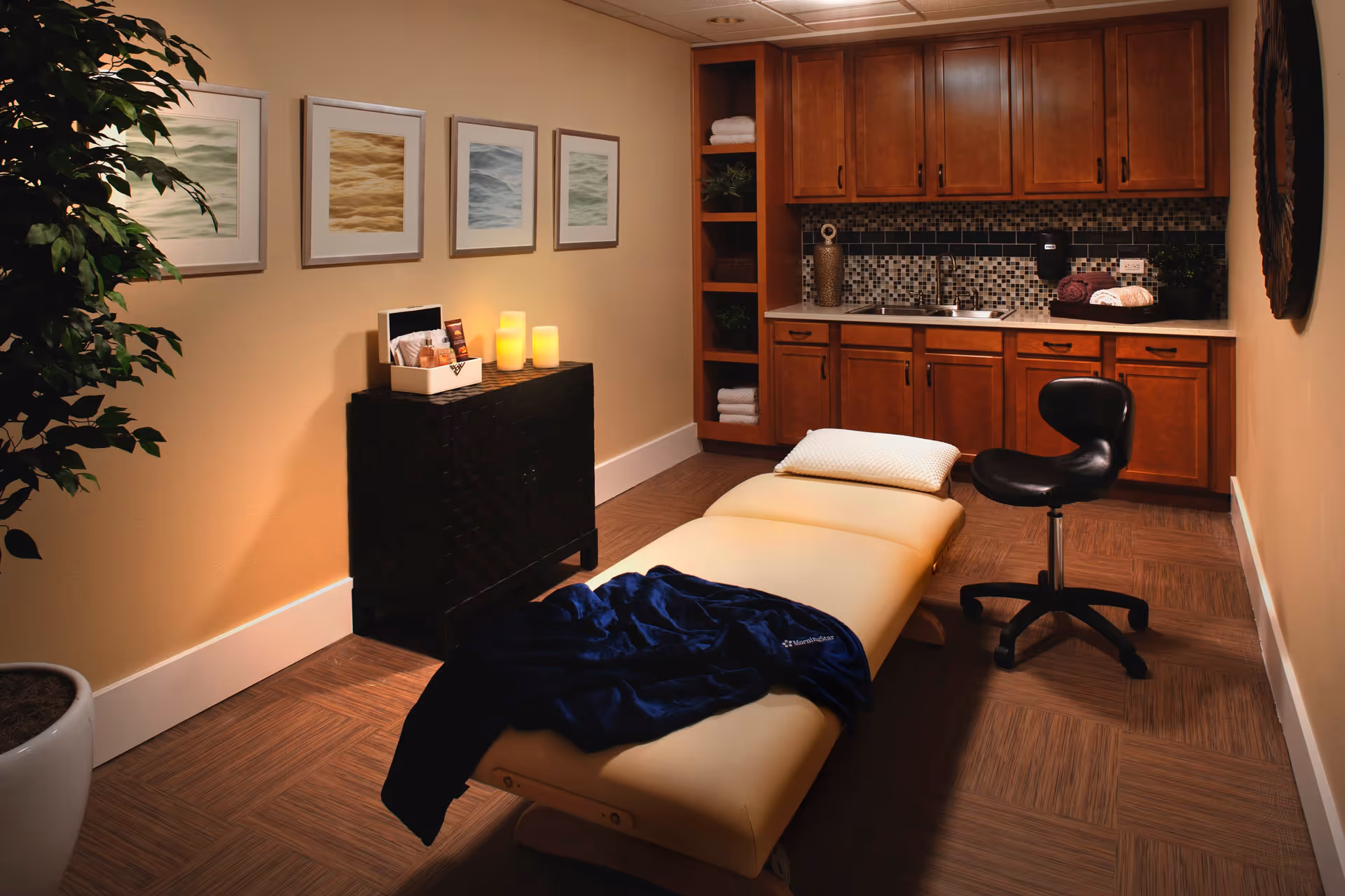 A cozy therapy or massage room with a padded treatment table covered partially by a dark blue blanket and a white pillow. There is a black rolling stool next to the table. The room has wooden cabinets with a mosaic tile backsplash, a countertop with a sink, and shelves holding towels and decorative items. On one wall, there are four framed abstract art pieces, and a small black cabinet with lit candles and a box of supplies. A large potted plant is in the corner, adding a touch of greenery.